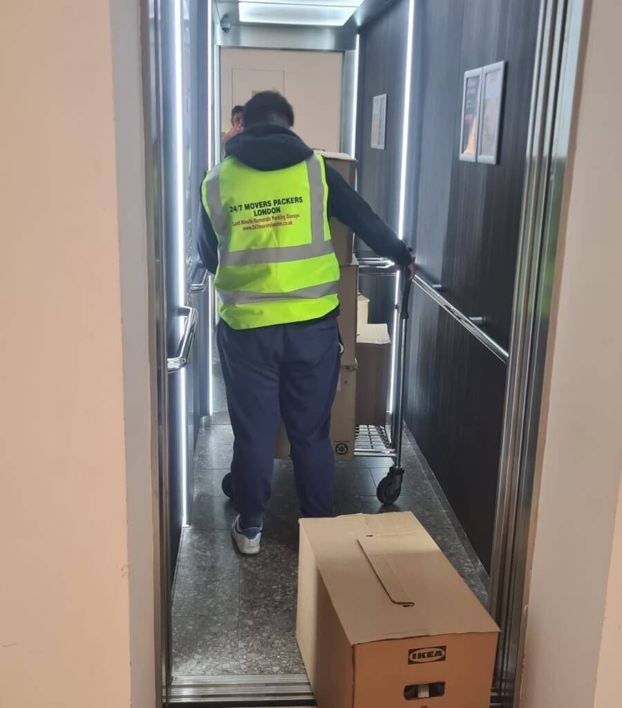 The worker of a moving company is wearing yellow shirt and carrying a trolley in which there are packed boxes.