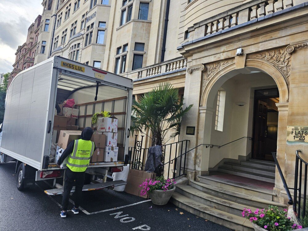A 24/7 Movers Packers London worker unloading packed boxes from a moving van outside a residential building with a stone entrance and potted flowers.