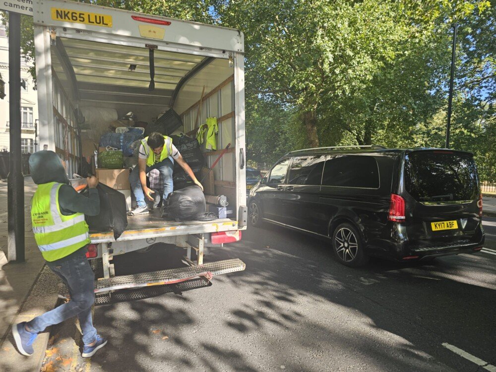 Movers wearing reflective vests unloading large black bags and boxes from a white truck parked on the roadside. A black van is parked nearby under the shade of tall trees.