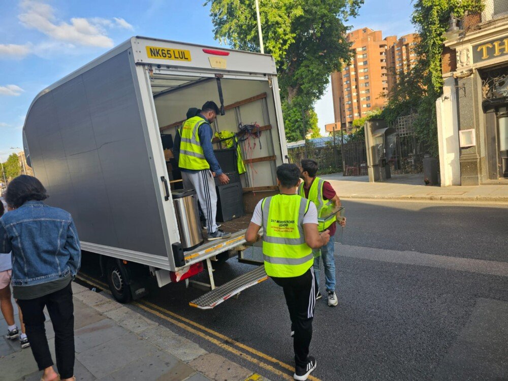 Moving team in reflective vests unloading furniture and equipment from the back of a white truck on a busy street. A man carries an item while another stands inside the truck arranging belongings.