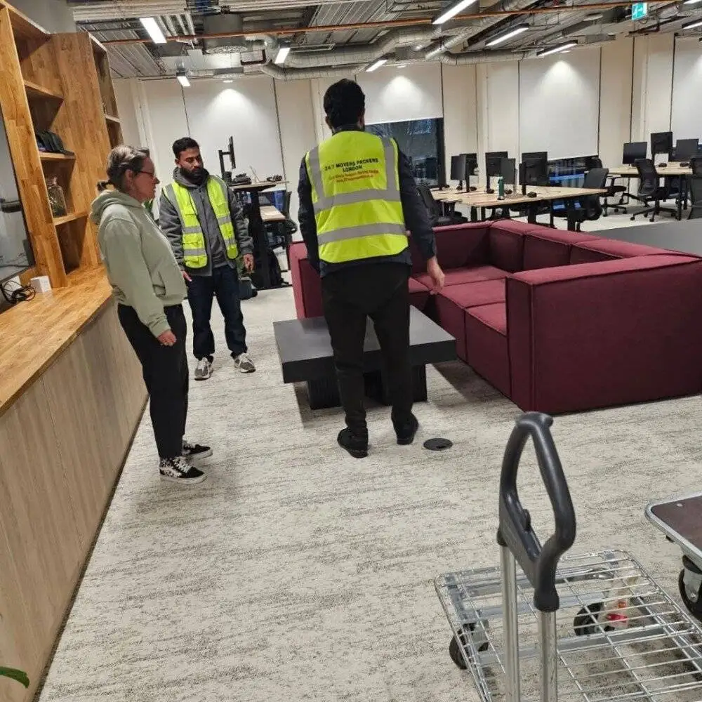 24/7 Movers Packers London team moving a maroon office sofa inside a modern workspace with desks and computers in the background.