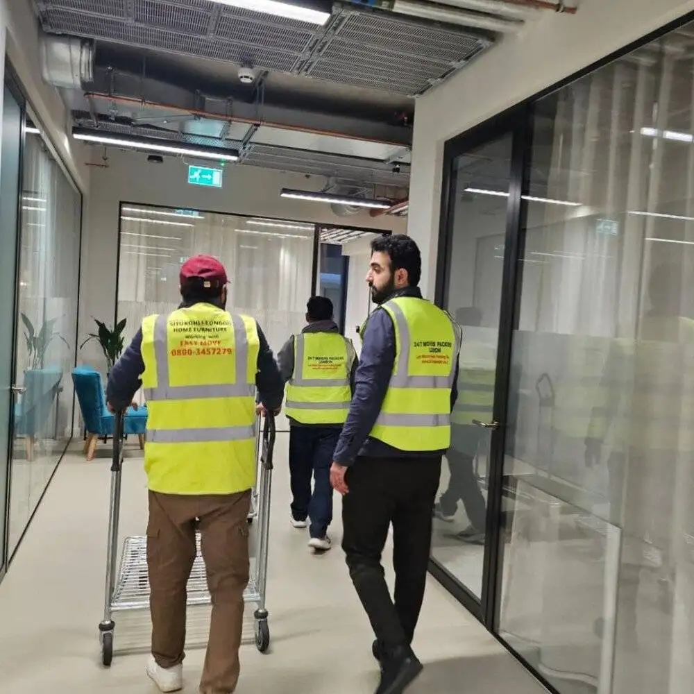 Movers wearing yellow safety vests are pushing trolleys with furniture down a modern office corridor.