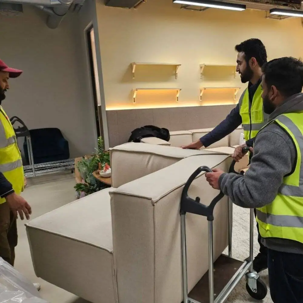 Three movers in high-visibility vests are positioning beige sofas onto a trolley inside an office space.