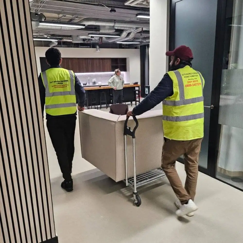 Two movers transport a beige sofa on a trolley through an office hallway towards a lounge area.