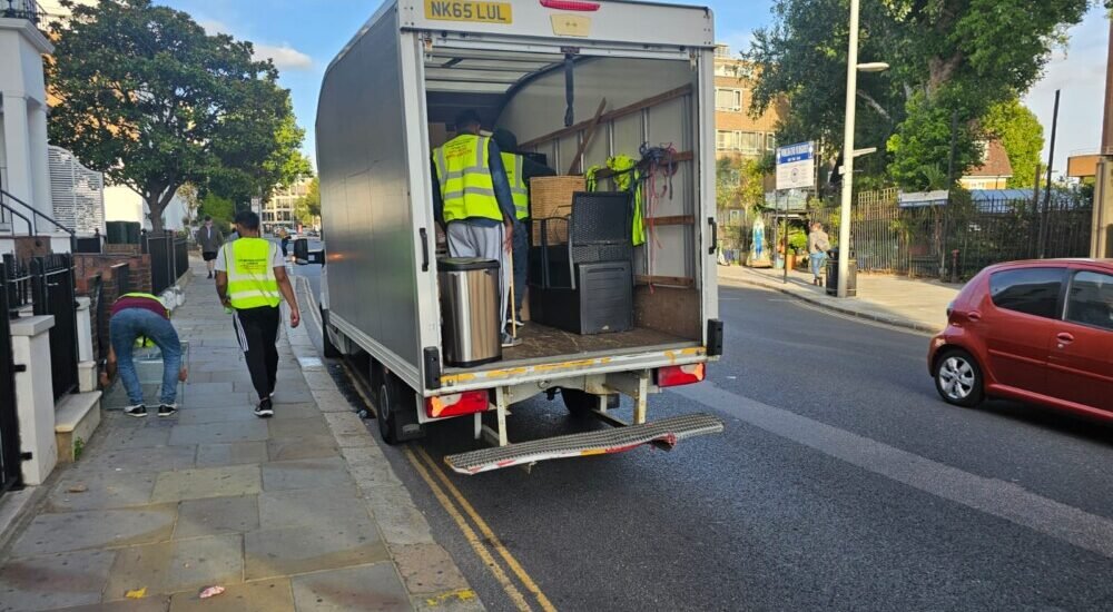 Workers in yellow reflective vests unloading furniture and items from a white moving truck parked on a city street. One worker bends down near the pavement while others assist with unloading.