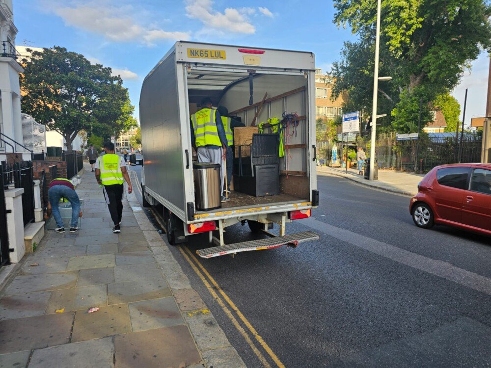 Workers in yellow reflective vests unloading furniture and items from a white moving truck parked on a city street. One worker bends down near the pavement while others assist with unloading.